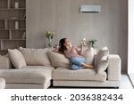 Relaxed young hispanic female homeowner sitting on huge comfortable couch, turning on air conditioner with remote controller, switching on cooler system, setting comfortable temperature in living room