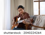 Creative hobby. Talented young mixed race female musician sit in armchair alone compose instrumental song using classic guitar. Smiling biracial lady play calm melody on musical instrument. Copy space