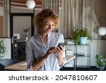 Happy young self employed woman using cellphone alone in office, reading text on screen, making mobile phone call, smiling, chatting online, getting good news, ordering, shopping via online app