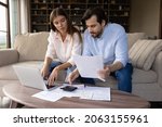 Small photo of Concentrated young family couple calculating expenditures, analyzing paper bank bills, managing monthly budget, planning investment, paying for service or utilities online using computer app.