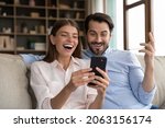 Small photo of Overjoyed young family couple looking at telephone screen, feeling excited getting shopping discount sale deal offer, celebrating reading online lottery gambling win notification, sitting on sofa.