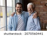Small photo of Warm relationship. Family portrait of mature father young adult son stand by window hug shoulders look at camera. Friendly aged grandfather grown grandkid feel emotional bonding enjoy pastime together