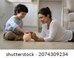 Happy Indian kid and young mom saving money together, putting cash into ceramic piggy bank. Mother playing with child on heating floor at home, teaching little son to invest money, planning future