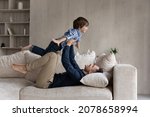 Small photo of Dad lying on couch lifts on outstretched hands little 6s son. Happy cute boy play plane fly in air while his father raise him up on arms. Active weekend at home, dreams about holiday, leisure concept