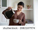 Smiling young indian ethnicity woman pouring hot water from kettle into cup, brewing tea or coffee, drinking beverage in modern kitchen, enjoying peaceful morning evening stress free carefree time.