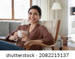 Small photo of Portrait of smiling beautiful indian ethnicity woman resting in cozy armchair with cup of coffee or tea in hands, enjoying carefree lazy relaxed morning weekend time lone in modern living room.