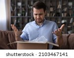 Small photo of Head shot unhappy dissatisfied man opening parcel at home, sitting on couch with cardboard box, angry displeased customer confused by wrong or damaged order, bad delivery shipping service concept