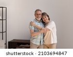 Small photo of Portrait of affectionate loving old woman cuddling shoulders of sincere mature husband, looking at camera posing in modern living room, feeling excited in own dwelling, retirement lifestyle concept.