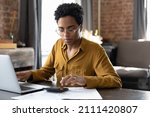 Focused millennial Black business woman calculating finance, money, using calculator, laptop computer at home workplace table, counting budget, paying bills, taxes, rent, mortgage fees