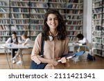 Small photo of Happy Hispanic gen Z student girl with headphones visiting public library for work on study research project, holding learning papers, notebook, looking at camera, smiling. Head shot portrait