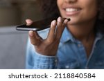 Happy teen African girl giving voice commands to virtual assistant on smartphone, speaking for recording audio message, holding, using mobile phone, smiling. Hand with cellphone close up