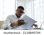 Serious African American manager in stylish glasses reading corporate document with note, sales report, boss order, doing paperwork. Legal expert checking document at workplace with laptop
