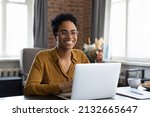 Joyful happy short haired Black business woman in glasses sitting at workplace with laptop in home office, looking at camera, smiling. Millennial worker, employee, entrepreneur head shot portrait