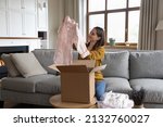 Small photo of Smiling woman unpack parcel box with delivered online fashion store clothes, check blouse quality, feel satisfied by received apparels. Client of e-commerce webstore, express delivery services concept