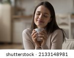 Close up beautiful woman relaxing on sofa with eyes closed, hold tea cup, take break at home enjoy favourite beverage looks carefree, relish aroma of coffee, daydreams alone indoor. No stress concept