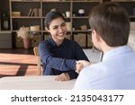 Small photo of Diverse gender entrepreneurs business partners sit at desk handshake close deal satisfied with negotiations result. Young Indian woman successful job applicant being hired on place shake man hr hand