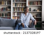Small photo of Calm middle-aged grey-haired woman sit on cozy fashionable sofa in living room staring into distance looks pensive relaxing alone at home. Carefree retirement, recollect past life, memories concept