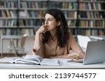 Young student girl sit at table with textbooks and laptop staring aside, studying alone in library, looks pensive and thoughtful search solution, prepare for exam, makes task feels confused or puzzled
