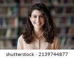 Small photo of Close up attractive brunette teenage girl with curly hair pose in library look at camera, head shot portrait on bookshelves background. Higher education institution student, excellent studies concept