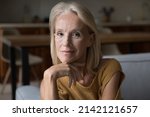 Smiling blonde middle aged 60s lady home head shot portrait. Skinny retired woman, pensioner, OAP sitting on couch, touching chin, looking at camera, posing for profile picture