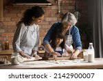 Small photo of Happy Hispanic mom and grandma teaching girl to bake, kneading, rolling dough for pastry together. Three family female generations cooking homemade bakery food, having fun at messy kitchen table