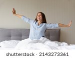 Happy pretty millennial woman awaking in good morning, feeling full of energy after sleep enough. Satisfied hotel guest sitting in beddings, on comfortable mattress, stretching hands