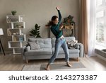 Small photo of Full of energy. Active young latin lady dance at living room alone do fitness exercise by live music from cellphone. Energetic joyful millennial woman renter tenant jumping having fun at home interior