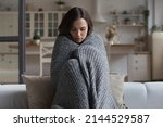 Stressed sad wrapped in plaid young woman feeling cold, ill, sick, suffering from fever, virus, trying to warm, sitting on couch at home. Frustrated shocked girl going through depression