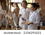 Small photo of Art school student and older teacher laugh during painting lesson. Indian woman holds palette, paintbrush stand near easel enjoy hobby, artistic practice and talk with tutor. Group art-class concept