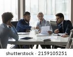 Small photo of Serious multiracial older and young businesspeople gathered in boardroom discuss financial statistics, analyze sales report, forecasting work together at office meeting. Teamwork, negotiations concept