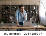 Work on idea. Thoughtful casual male in glasses stand by desk with pc look at distance touch chin create project ponder on business plan. Pensive young man scientist do research think on new solution