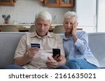 Shocked aged couple become victims of online fraud using credit card phone to pay for goods order service online on suspicious website. Frustrated older spouses overspending money at internet shopping
