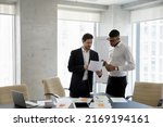 Small photo of Two multi ethnic businessmen speaking in office, consider documents, share information, planning collaborative project, working together in modern board room. Teamwork, workflow and business concept