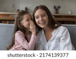 Small photo of Cute little daughter share secret, tells whispers gossips on young loving mothers ears, smile enjoy good friendly trustworthy relations sit on sofa at home. Understanding, family ties, trust concept