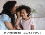 Small photo of Cheerful African girl listens mothers secret sit together on couch, enjoy trustworthy conversation, having friendly communication. Young mom whispering in ear, share gossips to cute smiling daughter