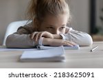 Small photo of Sad little Caucasian girl child sit on table at home distracted from studying lack motivation doing homework. Unhappy small kid feel lazy bored preparing task assignment. Boredom, education concept.