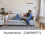 Small photo of Happy millennial couple of homeowners enjoying cool conditioned air, resting on couch together, using remote control for AC, cooling domestic equipment start. Home appliance concept