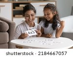 Small photo of Happy Indian daycare teacher showing flash cards to little kid, helping child with multiplication table study, giving math lesson, talking, smiling, laughing. Mom teaching girl to count