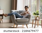 Joyful pretty millennial Indian cellphone user girl resting in armchair at cozy home, touching mobile phone screen, typing, chatting online, enjoying leisure, shopping on Internet, communication