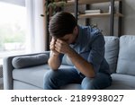 Sad depressed millennial man sitting on sofa in living room with head-down, suffers due to personal life troubles, goes through break up, split, divorce, after quarrel mood, feeling remorse concept