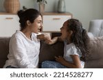 Small photo of Young Indian mother and cute daughter practicing speaking together, doing vocal exercises seated on sofa at home, therapist logopaedist correct pronunciation of preschooler girl, help to stutter kid