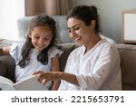 Small photo of Smiling Indian woman read a book to little daughter, preschooler 6s adorable girl sits on sofa enjoy mothers storytelling spend leisure together at home. Children development, hobby, good life habit