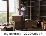 Small photo of Young man standing in unfurnished room near heap of cardboard boxes with personal belongings using smart phone during relocation day. Mobile app e-services for move-out, transporting company client