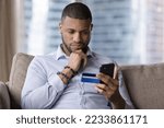 Small photo of Thoughtful African credit card user holding mobile phone, looking at screen, touching chin in doubts, making decision, using ecommerce banking app for making payment, shopping