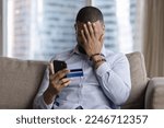 Small photo of Unsuccessful payment, internet scam, fraud victim. Upset African guy sit on sofa holds card and smartphone, cover face with palm feels shocked, overspending money, lost savings, money stolen from bank