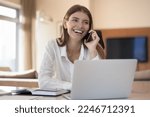 Small photo of Smiling 25s woman sit at desk with laptop and talks on smartphone, blab with friend, lead formal pleasant conversation, providing services to client using telecommunications mobile operator company