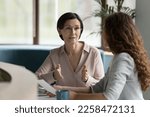 Confident mature business professional woman talking to younger female colleague at office table, speaking, gesturing, teaching, explaining work tasks. Elder mentor training intern