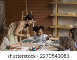 Small photo of Friendly young artistic class teacher helping pupil girl with drawing on canvas, discussing picture with African artist kid, smiling, laughing. Group of children learning painting in artistic school