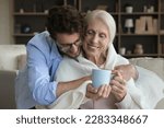 Small photo of Loving young adult son giving positive sick senior mother help, assist, care, wrapping mom shoulders into plaid, scarf, giving cup of hot drink, hugging, smiling, laughing