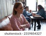 Unhappy abused young caucasian woman sitting on sofa in cafe separately from multiethnic diverse happy people, suffering from bullying, hurt by mockery gossip, unfair attitude or no friends support.